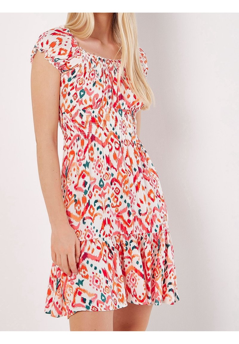 Femme portant une robe courte à motifs multicolores avec manches bouffantes, corsage froncé et ourlet volanté, posant devant un fond uni.