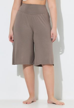 Plus-size person wearing loose, knee-length taupe shorts with a wide waistband, standing barefoot against a white wall.