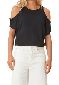 Femme portant un haut noir à épaules dénudées, associé à un pantalon blanc taille haute, se tenant avec les bras détendus le long du corps.