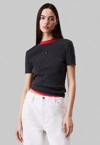 T-shirt a coste grigio scuro con colletto rotondo rosso, dotato di un logo sul petto. Abbinato a jeans bianchi a vita alta.