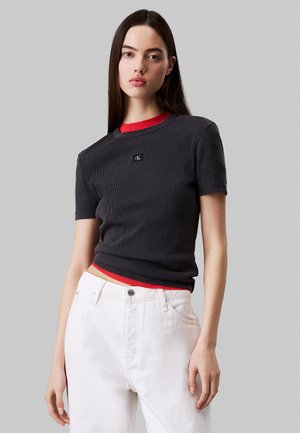 T-shirt a coste grigio scuro con colletto rotondo rosso, dotato di un logo sul petto. Abbinato a jeans bianchi a vita alta.