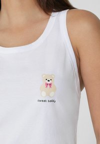 Camiseta sin mangas blanca con un dibujo de un oso de peluche de dibujos animados con un lazo rosa y el texto "dulce teddy" en negro debajo de la ilustración.