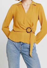 Donna che indossa una blusa a maniche lunghe giallo senape con dettaglio a anello in tartaruga legata in vita, abbinata a jeans in denim azzurro chiaro.