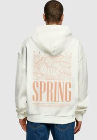 Overdimensioneret cremet hoodie med et pastelorange grafisk design med tropiske blade og ordet "SPRING" på ryggen. Blødt stof, afslappet pasform.