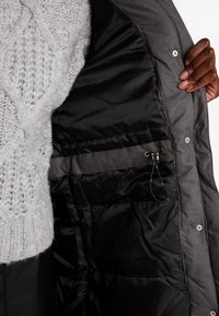 Giacca grigia in piuma con interno nero, caratterizzata da una trama trapuntata e un cordino elastico. Bottoni a pressione visibili e un morbido maglione grigio in maglia.