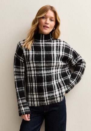 Donna con lunghi capelli biondi indossa un maglione a collo alto a quadri bianchi e neri e jeans scuri, in piedi davanti a una parete chiara e semplice.