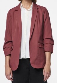 Blazer bordeaux avec un design ajusté, devant ouvert, deux poches et manches retroussées, associé à une chemise blanche en dessous.