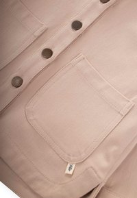 Camicia di corduroy beige con quattro bottoni in metallo, una tasca sul petto e un piccolo etichetta con un design a foglia sul bordo inferiore.