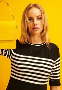 Keltainen nahkainen olkalaukku, jossa on läppä, tasaisen tekstuurin ja ompeluyksityiskohtien kanssa, jota kantaa malli musta-valkoisessa raidallisessa paidassa.