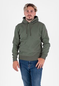 Felpa verde oliva con tasca a marsupio, coulisse regolabile, polsini a coste e orlo a coste. Indossata con jeans blu, con un'etichetta del logo sulla manica.