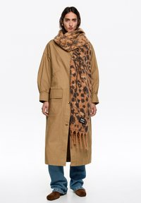 Beżowy długi płaszcz o luźnym kroju, z dwoma bocznymi kieszeniami, w połączeniu z brązowym szalem w cętki lamparta z frędzlami i metką marki.