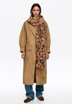 Beżowy długi płaszcz o luźnym kroju, z dwoma bocznymi kieszeniami, w połączeniu z brązowym szalem w cętki lamparta z frędzlami i metką marki.