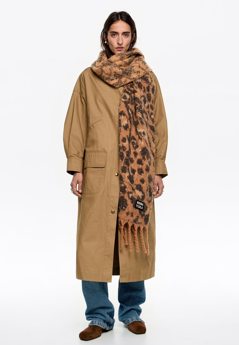 Beżowy długi płaszcz o luźnym kroju, z dwoma bocznymi kieszeniami, w połączeniu z brązowym szalem w cętki lamparta z frędzlami i metką marki.