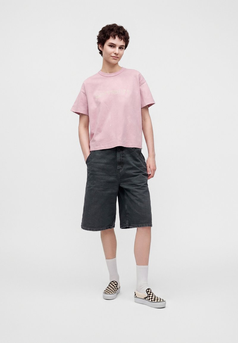 Junge Person mit kurzen lockigen Haaren, die ein ausgebleichtes pinkes T-Shirt, lange schwarze Shorts, weiße Socken und kariert Slip-On-Schuhe trägt und mit den Händen in den Taschen steht.