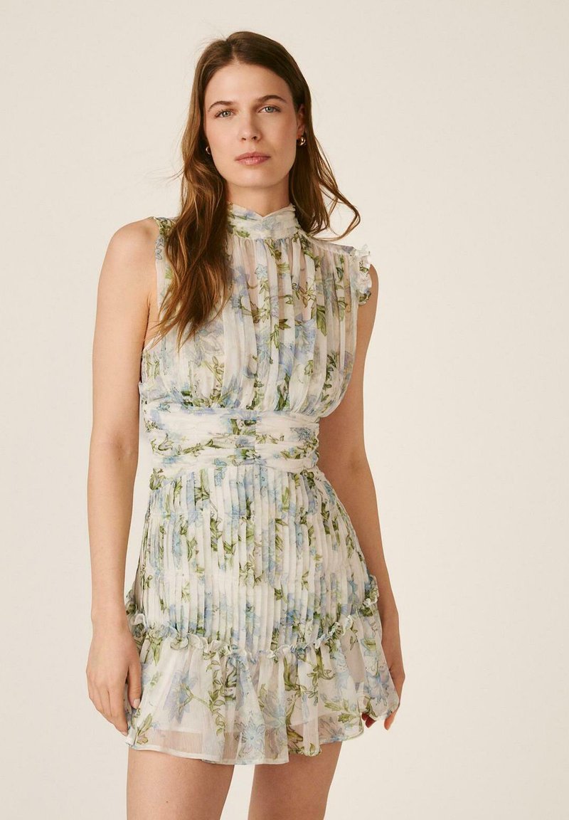 Mujer con vestido blanco plisado sin mangas con estampado floral y bajo con volantes, de pie frente a un fondo liso beige.