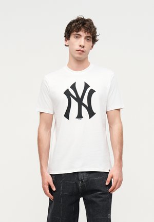 Junger Mann mit lockigem braunem Haar, der ein weißes T-Shirt mit einem großen schwarzen stilisierten NY-Logo und schwarze Denim-Jeans trägt.