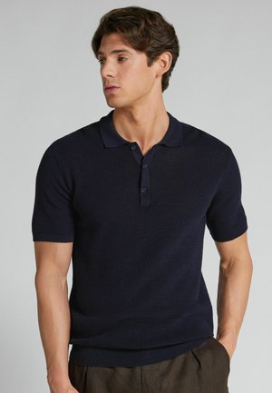 Jeune homme aux cheveux bruns portant un polo tricoté à manches courtes bleu marine foncé et un pantalon marron, regardant vers sa gauche sur un fond uni.