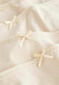 Lichtbeige stof met delicate kantafwerking en kleine crèmekleurige strikjes op regelmatige afstand. Zachte textuur; het ontwerp bevat golvende randen en subtiele details.