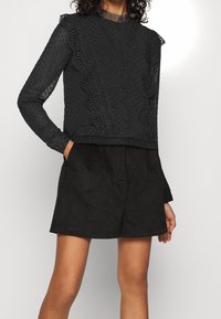 Musta paita, jossa on rakenteellisia pisteitä, pitsiaksentteja ja röyhelöreunat. Pitkät hihat ja korkea kaulus. Yhdistetty mustiin shortsihin.