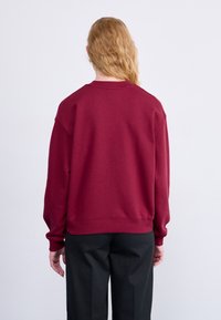 Sweat-shirt bordeaux avec un col rond classique et des manches longues, présentant une coupe décontractée et des poignets côtelés. Matière en mélange de coton lisse.