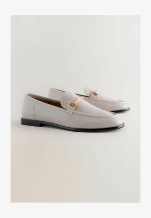 Helle graue Lederloafer mit spitzem Zehenbereich, versehen mit einem goldfarbigen Metallakzent und einem niedrigen schwarzen Absatz. Glatte Textur, minimalistisches Design.