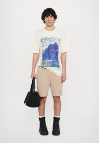 Creme-farbendes Graphic-T-Shirt mit blautem Druck, der die Architektur von Istanbul zeigt, kombiniert mit beigen Shorts und schwarzen Reißverschlussstiefeln. Schwarze Tasche in der Hand.