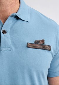 Vaaleansininen polo-paita, jossa on teksturoitu kangas. Paita sisältää kahden napin listan ja rintataskun, johon on ommeltu "International Speedway" -merkki.