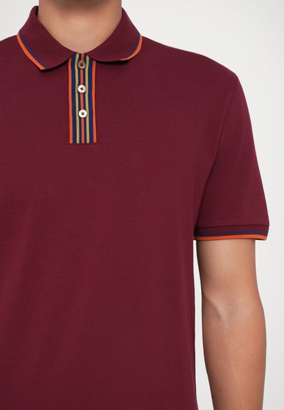 Μπορντό πόλο μπλούζα με πλαίσιο δύο κουμπιών, διαθέτοντας ριγέ γιακά και μανσέτες σε πορτοκαλί, μπλε και κίτρινες λεπτομέρειες.