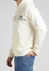 Sudadera blanca de algodón con bolsillo frontal, capucha con cordón y logo "Lee" bordado en marrón. Puños y dobladillo acanalados, corte relajado.