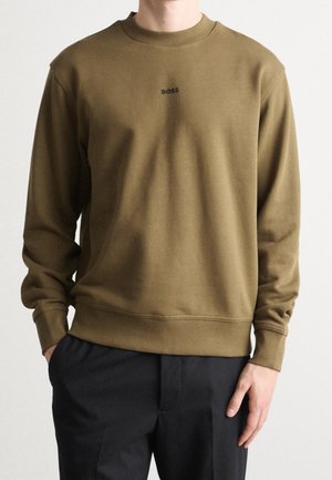 Persoon draagt olijfgroene trui met ronde hals en klein zwart "BOSS" logo op de borst en zwarte broek, staand tegen een effen achtergrond.
