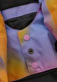 Färgglad tyg med en gradient från orange till lila, med ett mönstrat struktur. Två knappar: en rosa, en svart, vid öppningen.