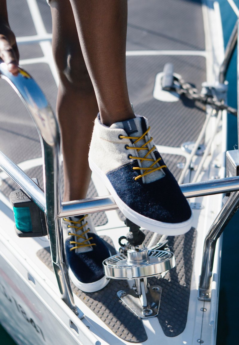 Buty żeglarskie w odcieniach granatu i szarości, z teksturowanym materiałem, białą gumową podeszwą i żółtymi sznurówkami, umieszczone na metalowej balustradzie.
