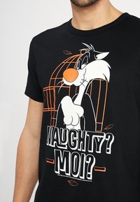 Hombre vestido con una camiseta negra que presenta al personaje de dibujos animados Sylvester el gato y el texto "¿Travieso? ¿Yo?" en blanco y naranja.