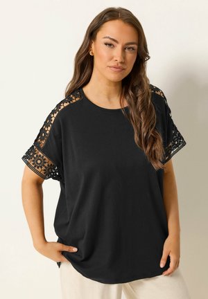 Kobieta ubrana w czarną bluzkę z kwiatową koronką na ramionach i rękawach, w połączeniu z jasnymi spodniami, stoi na jednolitym tle.