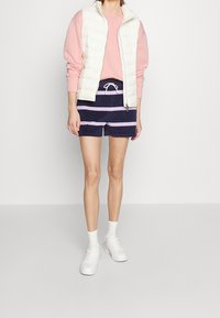 Gilet en duvet blanc sur un sweat-shirt rose, associé à un short marine avec des rayures roses horizontales, des baskets blanches et des chaussettes à la cheville.