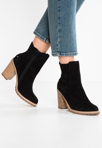Botines de ante negros con un tacón de madera texturizado, cremallera lateral y paneles elásticos, combinados con jeans azules recortados.