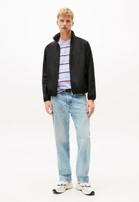 Zwarte bomberjack met hoge kraag, gedragen over een multicolor gestreepte T-shirt. Lichtblauwe straight-leg jeans en witte sneakers maken de outfit compleet.