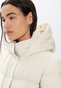 Krémová péřová bunda s vysokým límcem a nastavitelnou kapucí. Měkká textura, s kapsou na zip a minimálními švy.