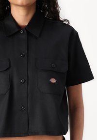Camisa preta de manga curta feita de tecido suave, com dois bolsos no peito e um pequeno emblema no bolso esquerdo. Frente com botões.