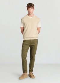 Pull sans manches beige en tricot porté sur un t-shirt crème, associé à un pantalon taillé vert olive et des chaussures en daim camel.