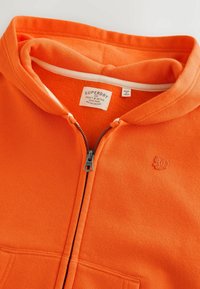 Sweat à capuche zippé orange avec doublure en polaire, bordure intérieure blanche au col, logo Superdry sur la poitrine et étiquettes de la marque à l'intérieur de l'encolure.