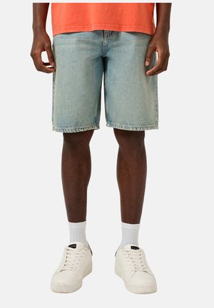 Homme portant un short en jean bleu clair, des chaussettes blanches, des baskets blanches et une chemise orange, les mains détendues le long du corps.