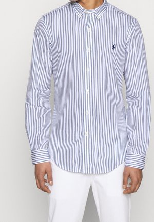 Mężczyzna noszący białą koszulę zapinaną na guziki z cienkimi pionowymi niebieskimi paskami oraz małym haftowanym logo konia na lewej piersi, w połączeniu z białymi spodniami.