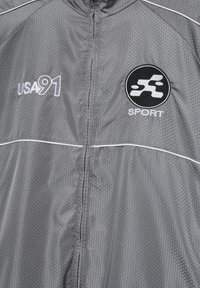 Sivý športový kabát s hexagonálnym vzorom, predný zips, text "USA 91" na ľavej strane hrudníka a kruhové logo "SPORT" na pravej strane hrudníka.