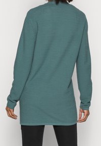 Persona vistiendo una túnica de manga larga acanalada en color verde azulado, vista desde atrás, combinada con pantalones negros, frente a un fondo claro y liso.