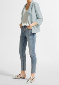 Ljusblå kavaj över en vit topp, kombinerad med figursydda ljusblå jeans och silverfärgade remsandaler. Ensemblet har en avslappnad, skräddarsydd stil.