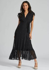 Robe maxi noire avec un design enveloppant, des manches à volants, des détails brodés et un ourlet transparent. Portée avec des sandales à plateforme beiges.