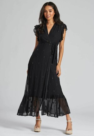 Abito maxi nero con design avvolgente, maniche a volant, dettagli ricamati e orlo trasparente. Indossato con sandali con pianta alta color beige.