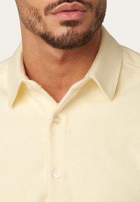 Gelbes, strukturiertes Stoffhemd mit einem Standardkragen, das sichtbare Knöpfe und ein leicht strukturiertes Design aufweist. Nahaufnahme des Kragens.