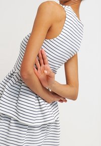 Proužkované šaty bez rukávů v bílé a námořnické modré barvě, s přiléhavým živůtkem a rozšířenou peplum sukní, vyrobené z hladké, lehké látky.